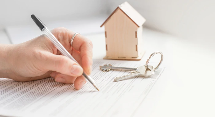 Person signing a document with a model house and keys on the table, symbolizing a real estate transaction.