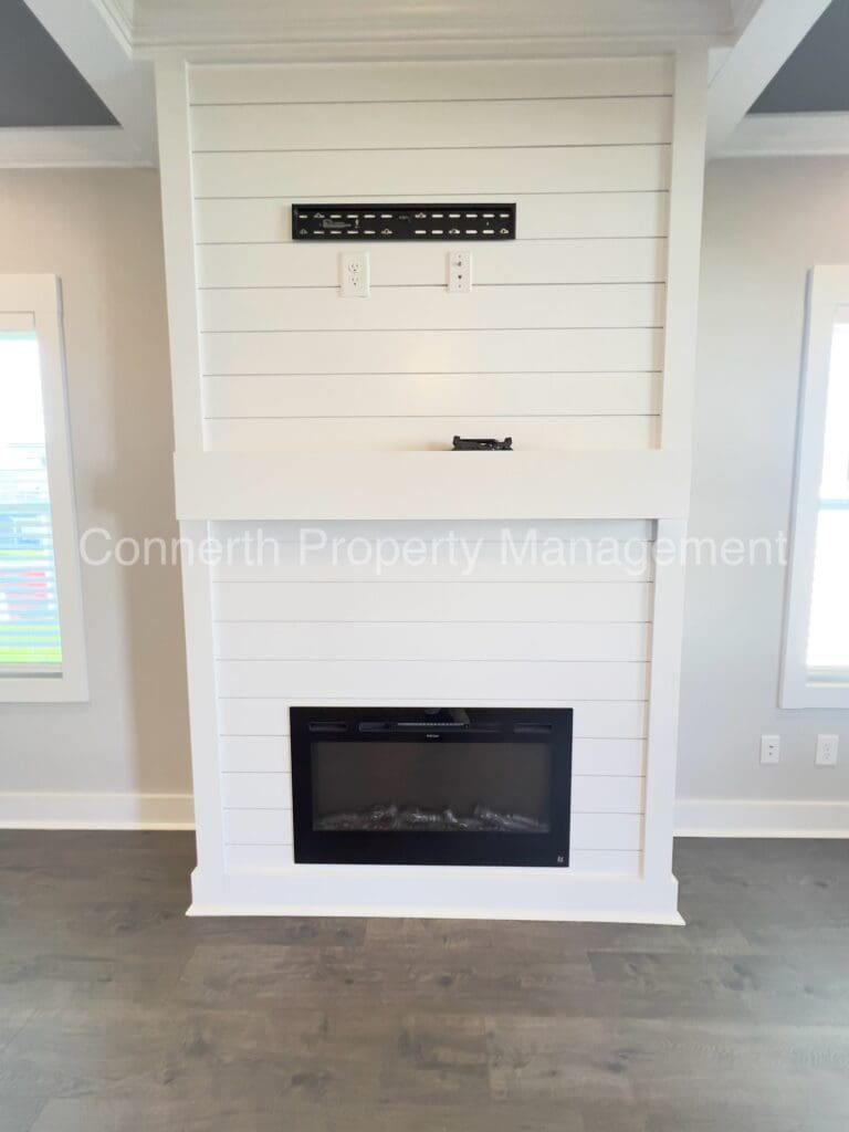 Modern electric fireplace built into a white columned structure with a decorative upper panel and a black mantel, set against a grey floor and neutral walls.