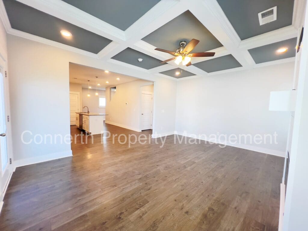 Empty living room with coffered ceiling, hardwood floors, and a ceiling fan, leading into an open kitchen.
