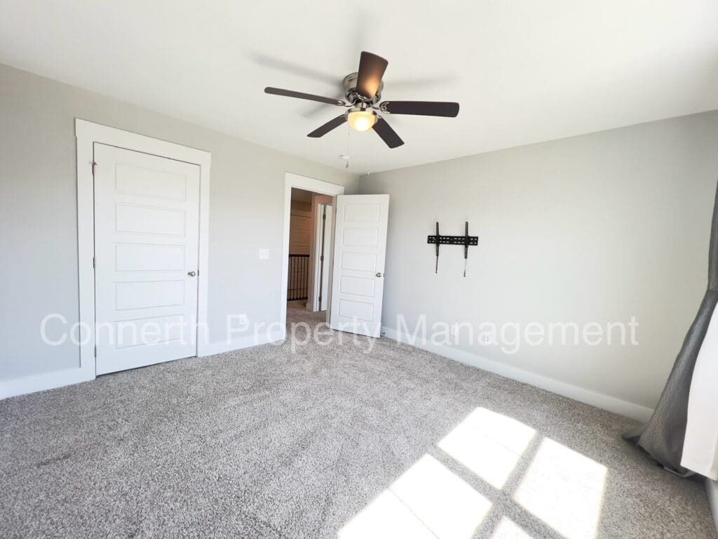 Empty residential room with gray carpet, white walls, a ceiling fan, a mounted tv bracket, and three doors. sunlight streams through a window, casting light patterns on the floor.