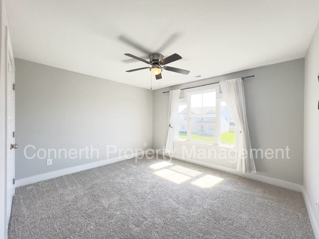 Empty room with gray walls, carpeted floor, ceiling fan with light, and large window draped with sheer curtains.