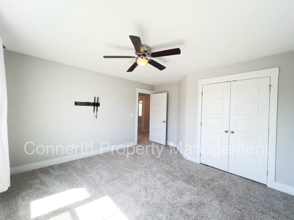 Empty room with carpeted floor, ceiling fan with light, two closed white doors, and a wall-mounted tv bracket.