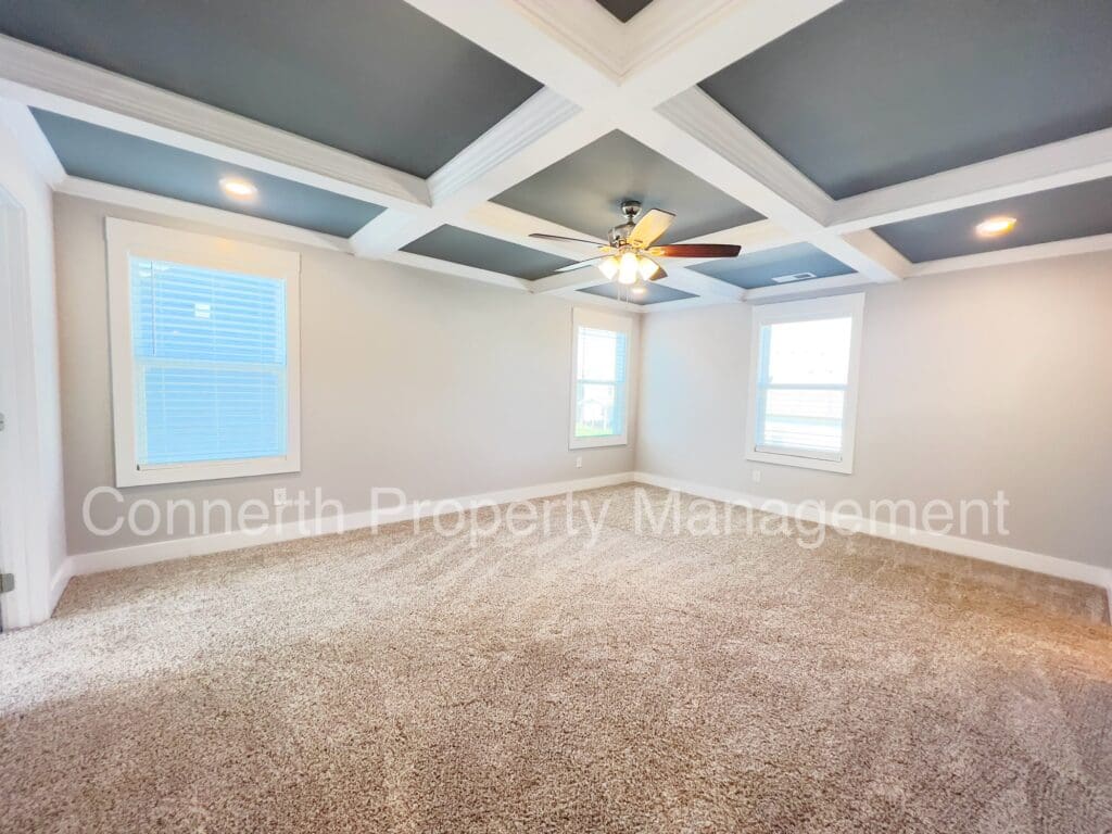 Empty living room featuring beige carpet, gray walls, a coffered ceiling with a fan, and natural light coming through windows.