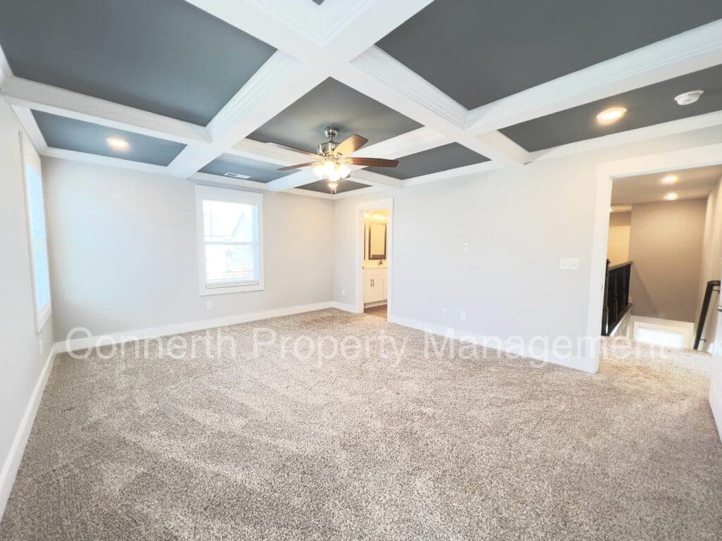 Empty living room featuring beige carpet, gray walls, a coffered ceiling with a fan, and natural light coming through windows.