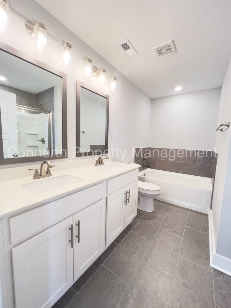 Modern bathroom with a double sink vanity, large mirrors, a bathtub, and a separate toilet area, featuring gray floor tiles and white walls.