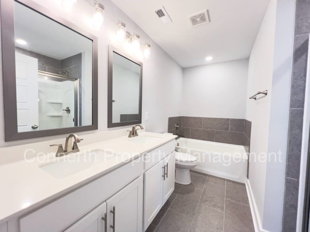 Modern bathroom with double vanity, large mirrors, white cabinets, gray walls, and a toilet visible in a mirrored reflection.
