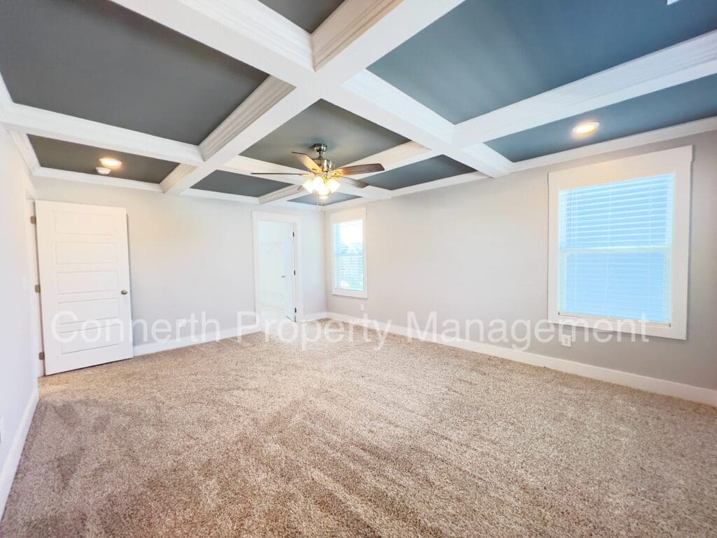 Empty residential room with textured carpet, coffered ceiling, ceiling fan, and two windows with blinds. white door on left, watermark by connerty property management.