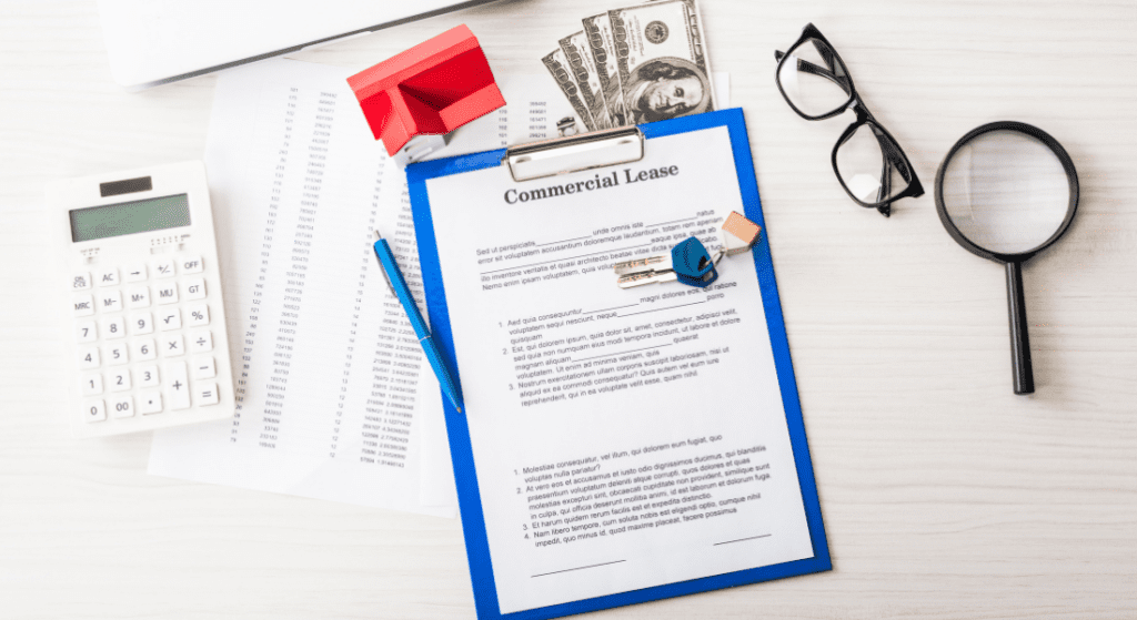 A commercial lease agreement on a clipboard is surrounded by a calculator, office supplies, glasses, a magnifying glass, keys, cash, and financial documents on a desk.