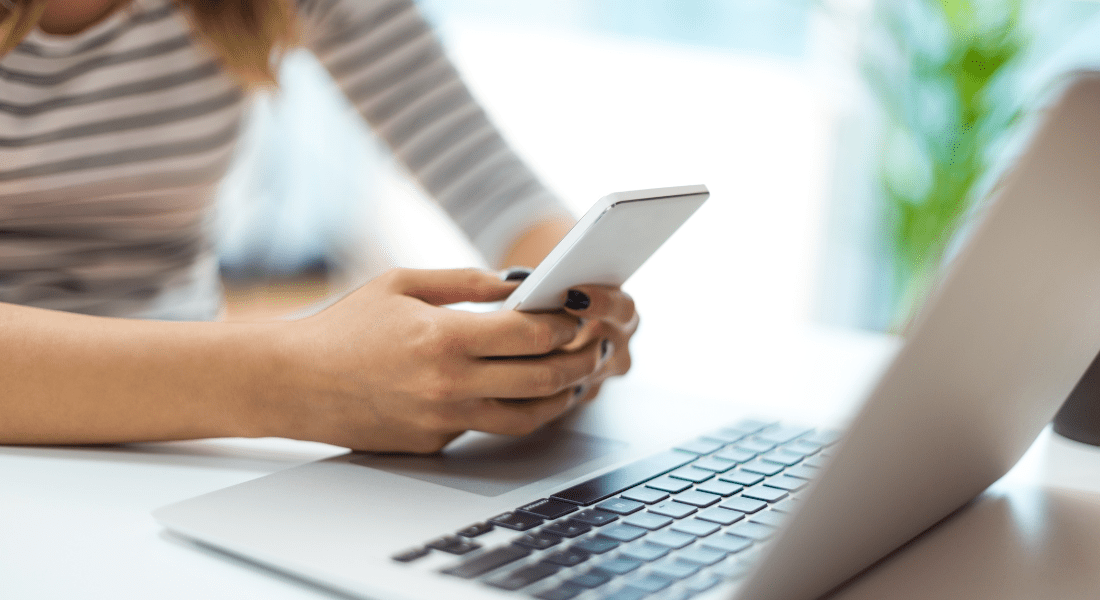 A person uses a smartphone while seated at a desk with an open laptop. Importance of Tenant Web Access in Property Management