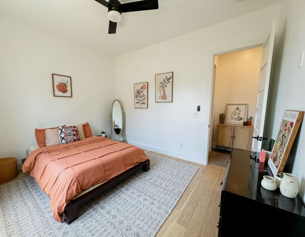 Bedroom with a queen-sized bed, peach bedding, and framed artwork on white walls. There is a black dresser, a wall-mounted mirror, and a ceiling fan. An open door reveals another room with a small cabinet.