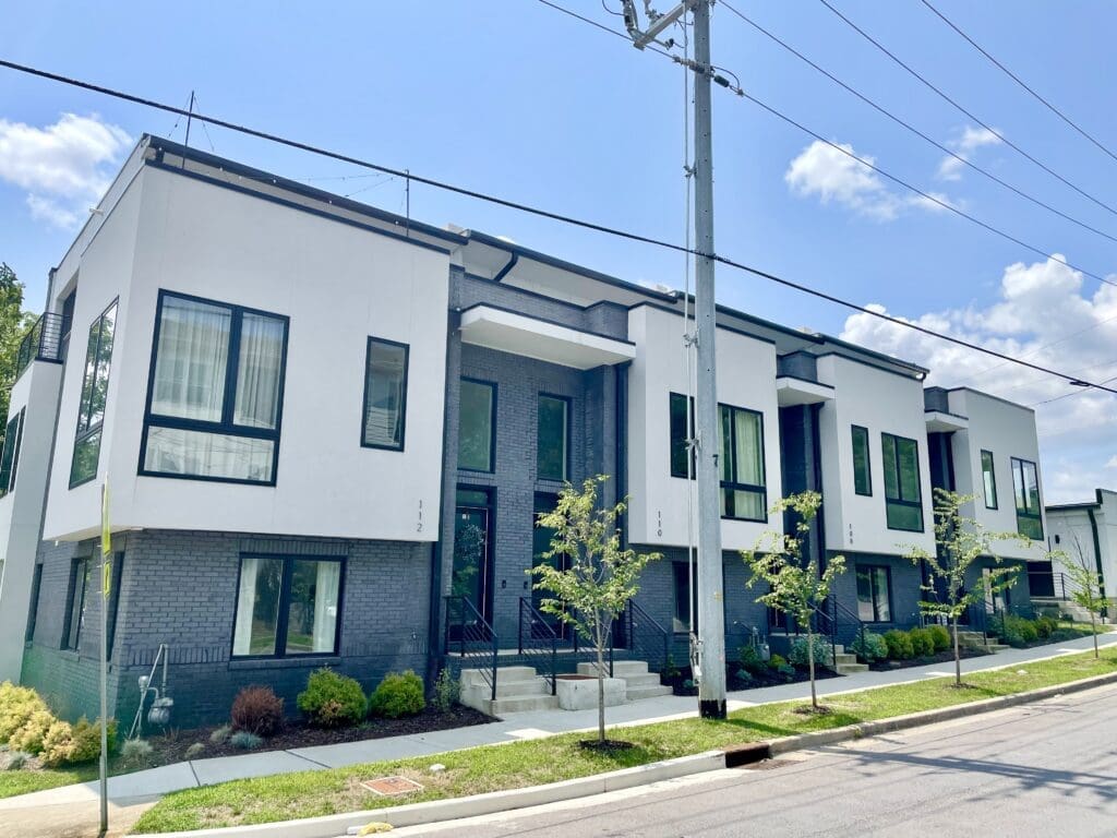 Modern, two-story apartment building with gray brick and white accents, large windows, a sidewalk in front, small trees, and power lines overhead under a clear sky.