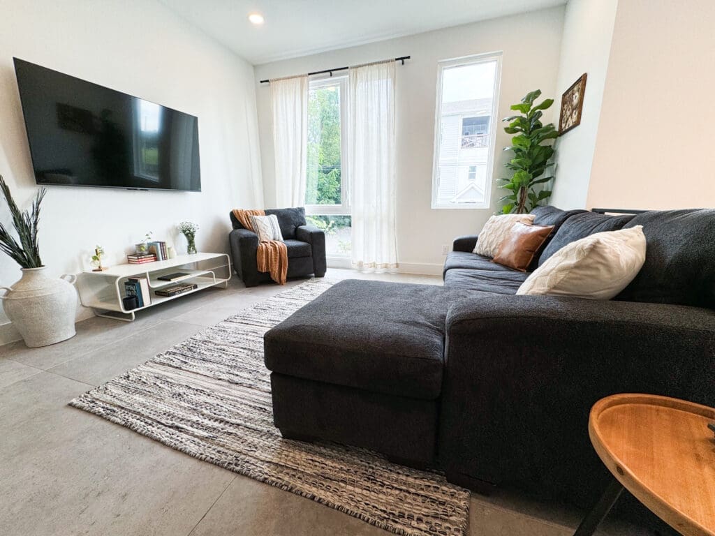 A modern living room with a large black sectional sofa, a wall-mounted TV, a striped rug, a small wooden side table, potted plants, and a window with sheer curtains.