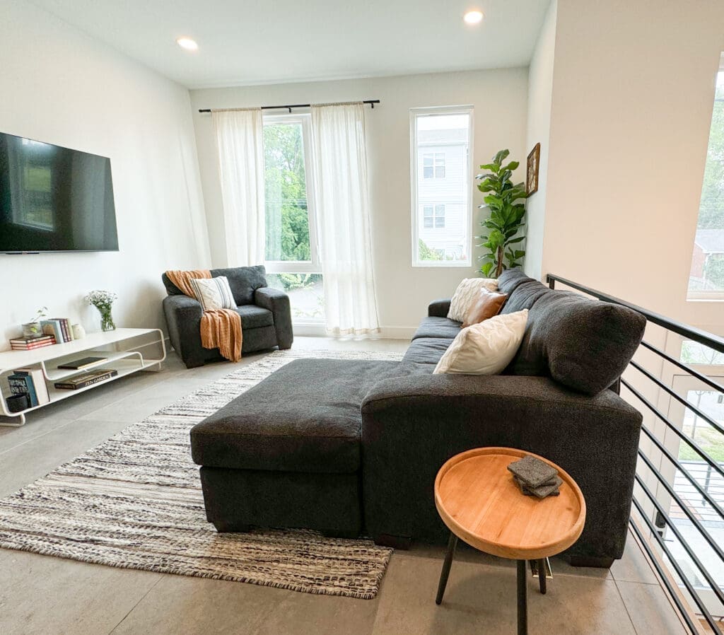 A modern living room with a dark sectional sofa, a wooden side table, a flat-screen TV on the wall, a large window, a striped rug, and a potted plant in the corner.