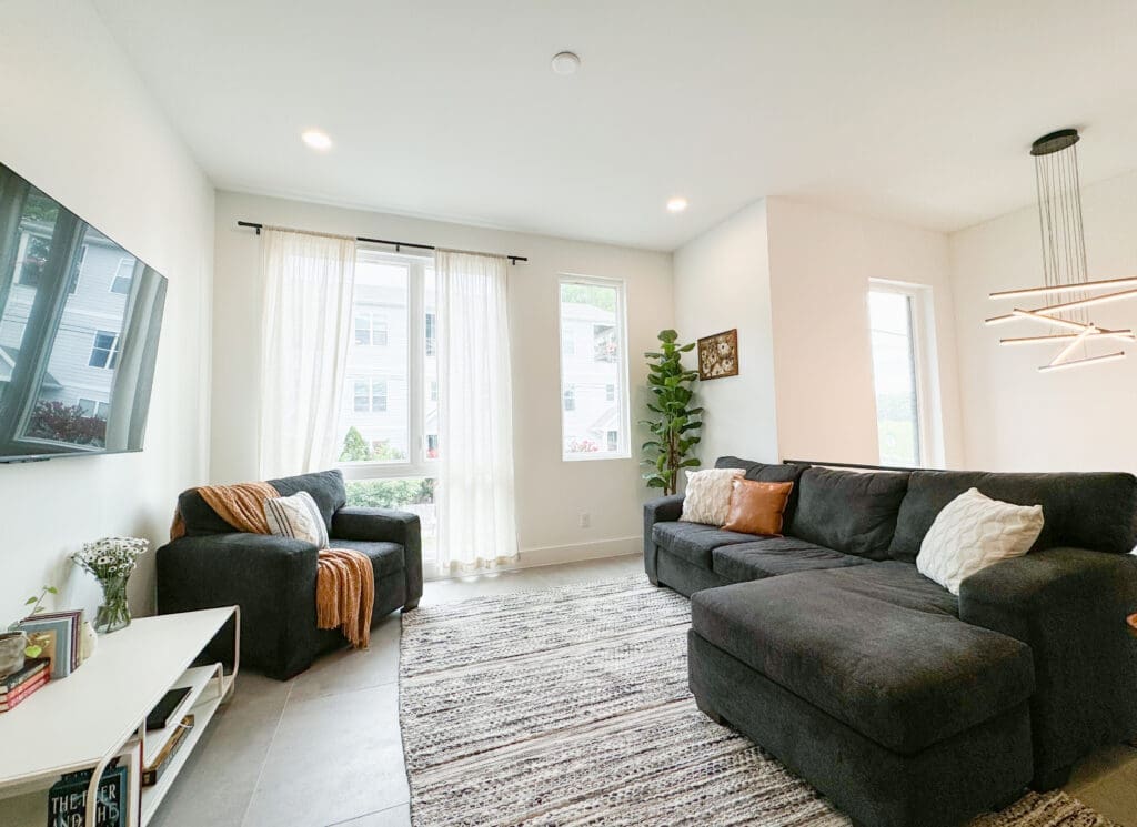 A modern, bright living room with a large sectional sofa, a single armchair, a mounted flat-screen TV, and a potted plant. Natural light comes through two large windows with sheer curtains.