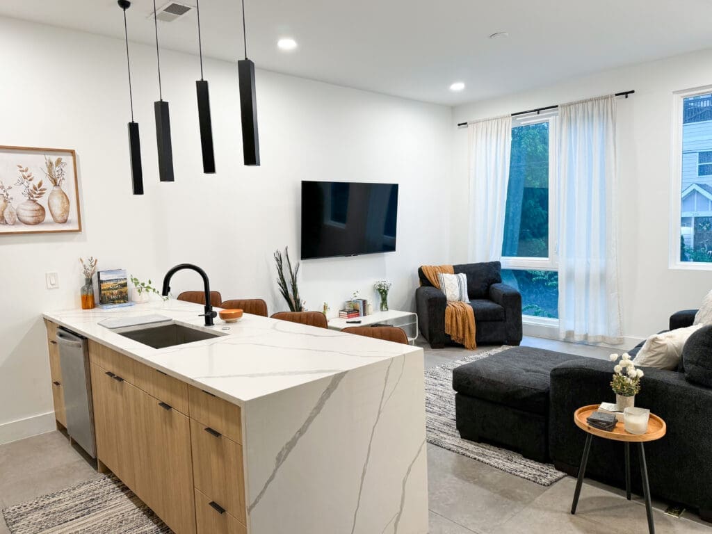 Modern living room with a kitchen island in the foreground, equipped with a sink. The room features a wall-mounted TV, a sofa, chairs, and large windows with white curtains. Black pendant lights hang above the island.