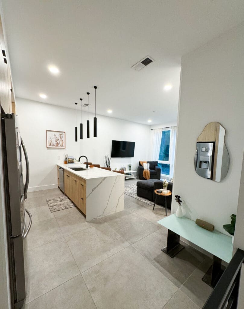 Modern kitchen with beige cabinets, island with marble countertop and black pendant lights. Adjacent living room with a wall-mounted TV, black sofa, and large window.