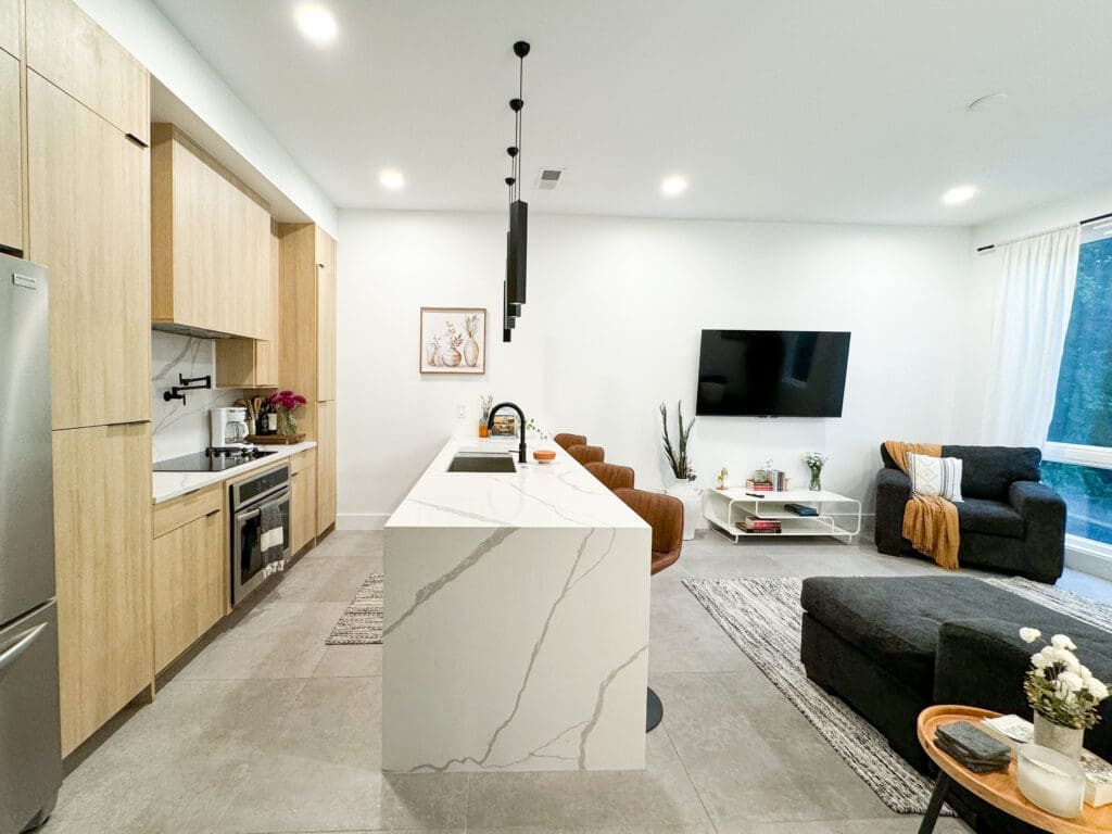 A modern open-concept kitchen and living area with light wood cabinetry, a white marble island, a wall-mounted TV, a gray sectional sofa, an armchair, and minimalistic decor.