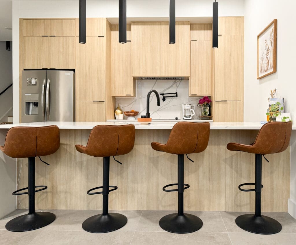 A modern kitchen with light wood cabinetry, a stainless steel refrigerator, a white countertop, and four brown leather barstools at the island.