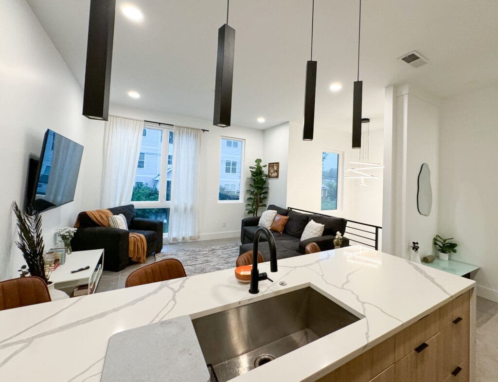 Modern open-concept living room and kitchen with a large island, pendant lighting, a wall-mounted TV, and a seating area featuring a sofa, armchairs, and a coffee table near a window.