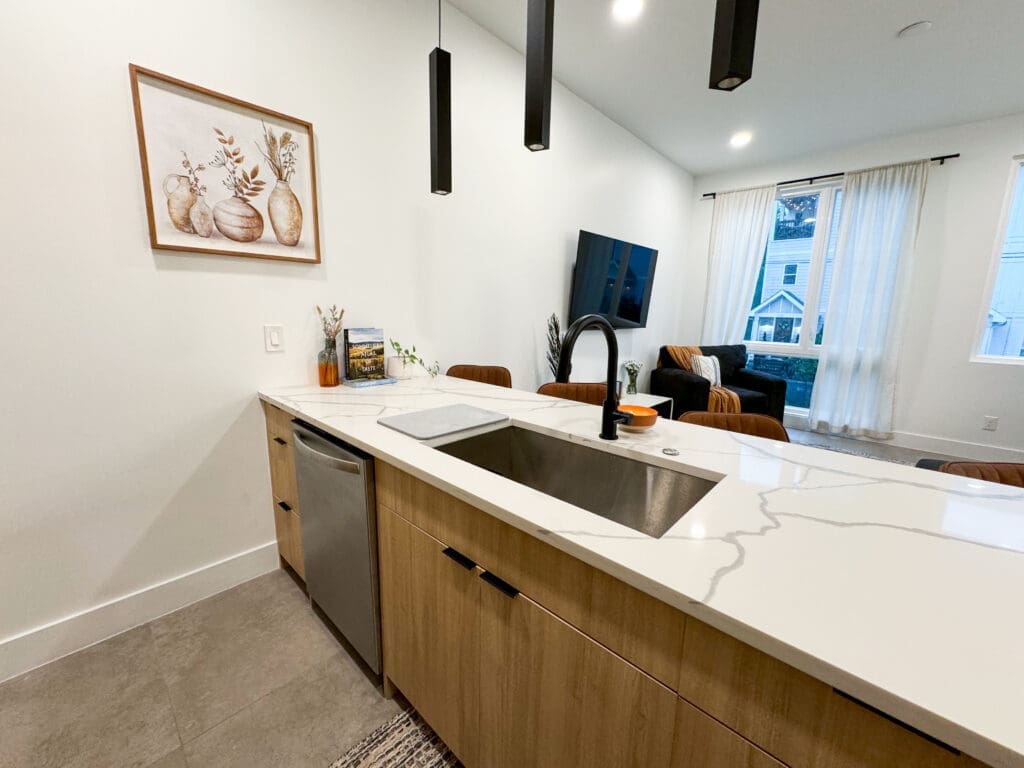 Modern kitchen with a center island featuring a sink and faucet. The island has wooden cabinets and a marble countertop. In the background, a living area with a wall-mounted TV and window with curtains is visible.