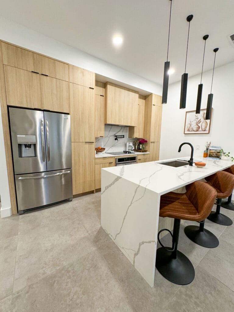 Modern kitchen with light wood cabinets, stainless steel refrigerator, built-in oven, and white marble countertops. An island with integrated sink and three bar stools is centrally located.