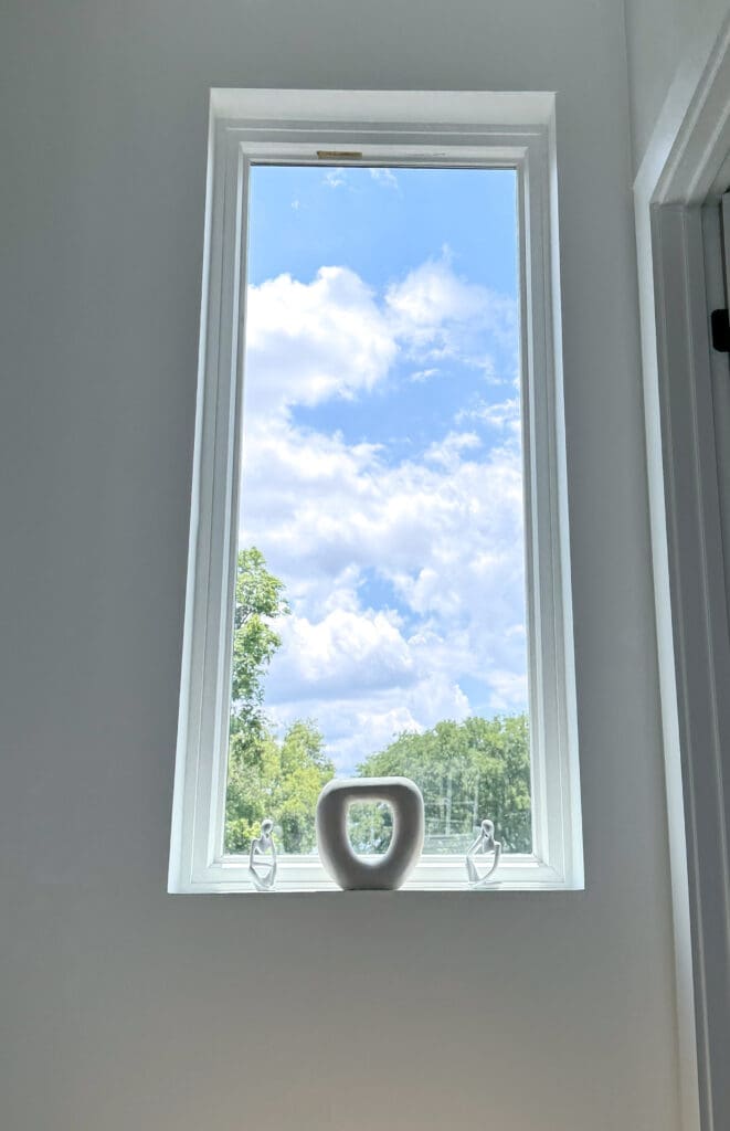 A tall window with a white frame reveals a clear blue sky with fluffy clouds and a view of green trees. A white ceramic vase sits on the window sill.