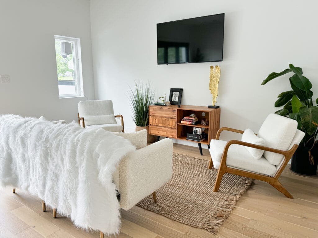 A modern living room with white armchairs, a couch draped with a fluffy blanket, a wooden TV stand, wall-mounted TV, potted plants, a rug, and a window allowing natural light to enter.