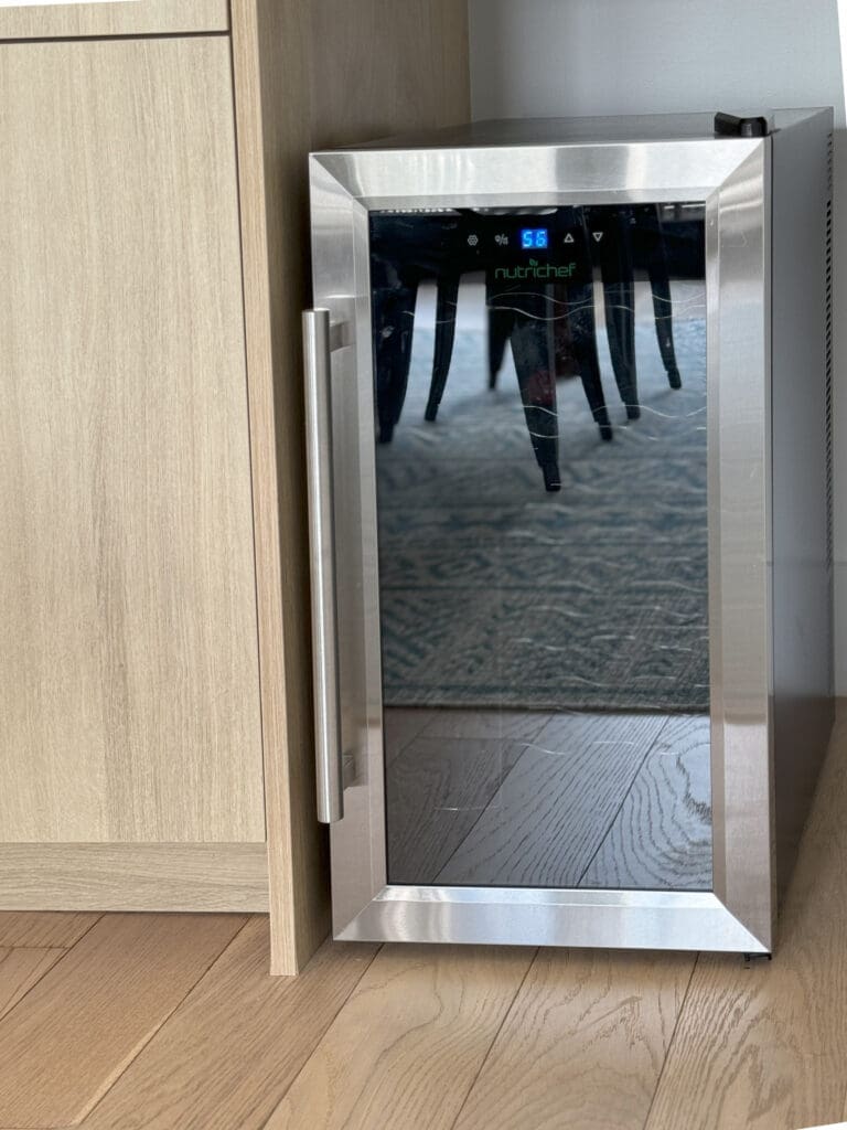 A stainless steel wine cooler with a digital temperature display showing 55 degrees Fahrenheit, partially reflected in its glass door. The cooler is placed next to a wooden cabinet.