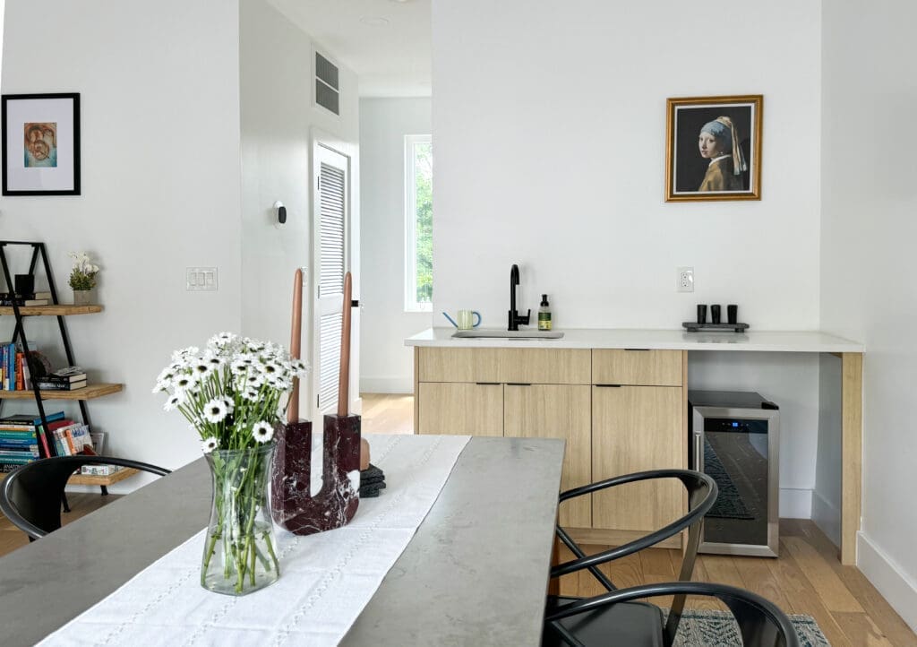 Modern kitchen with light wood cabinetry, a small refrigerator, and a countertop. A vase with white flowers and a decorative item are on the dining table. Framed artwork is hung on white walls.