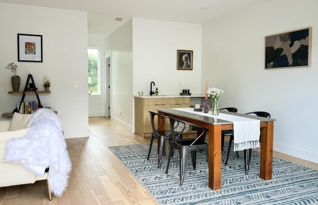 A modern dining area with a wooden table, black chairs, and a decorative runner. A white wall features paintings and a doorway leads to a sink area. A living area with a bookshelf is on the left.