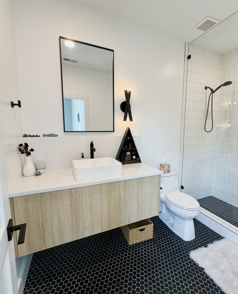 Modern bathroom with a wall-mounted wooden vanity, vessel sink, square mirror, black sconce light, and walk-in shower with white tiles and black fixtures. Black hexagonal floor tiles.