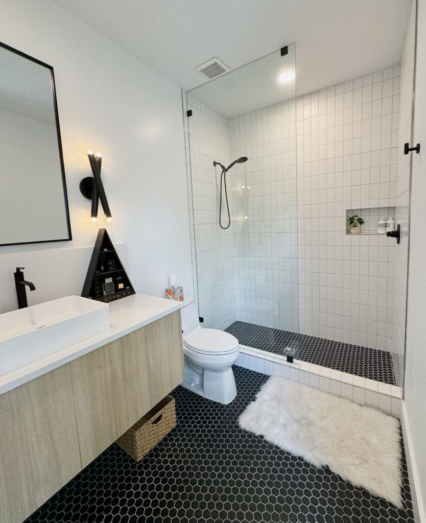 Modern bathroom with a glass-enclosed shower, white square tiles, black plumbing fixtures, a white vanity with a wooden base, a wall-mounted mirror, a black hexagonal tile floor, and a fluffy white rug.