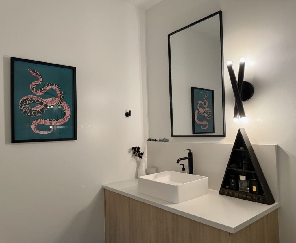 A modern bathroom with a white sink, wooden vanity, and a triangular shelf with toiletries. A framed artwork of a snake is on the wall, and a rectangular mirror is mounted above the sink.