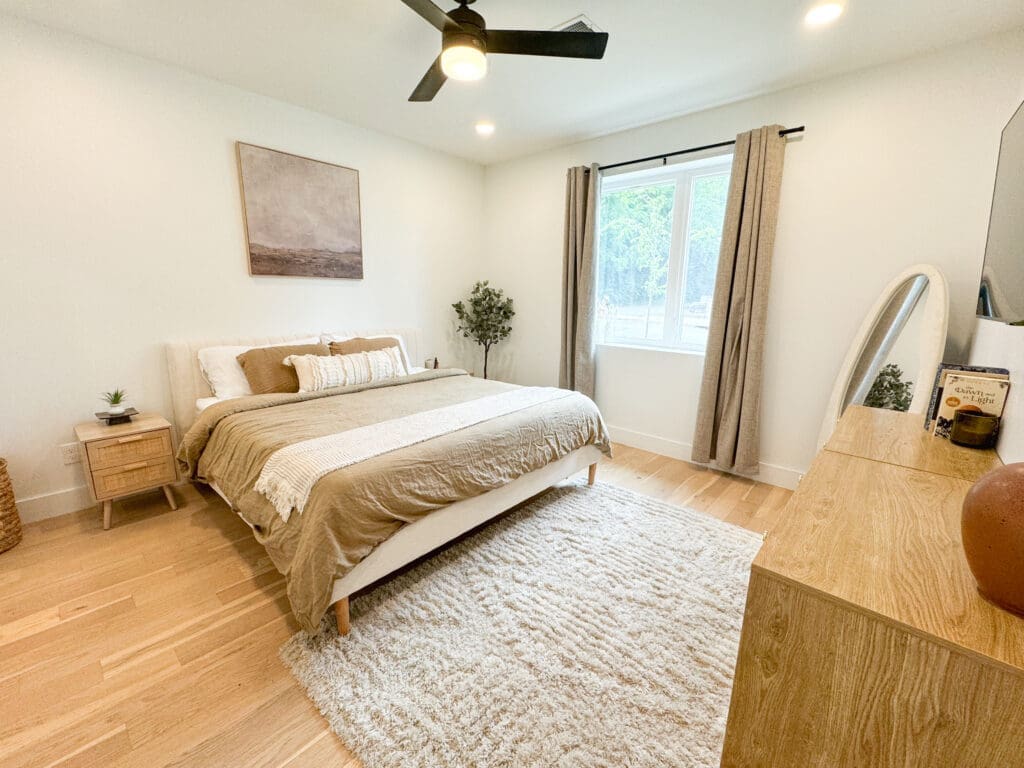 A minimalist bedroom featuring a neatly made bed with beige linens, two wooden nightstands, a dresser, a large window with curtains, a rug, and a ceiling fan.