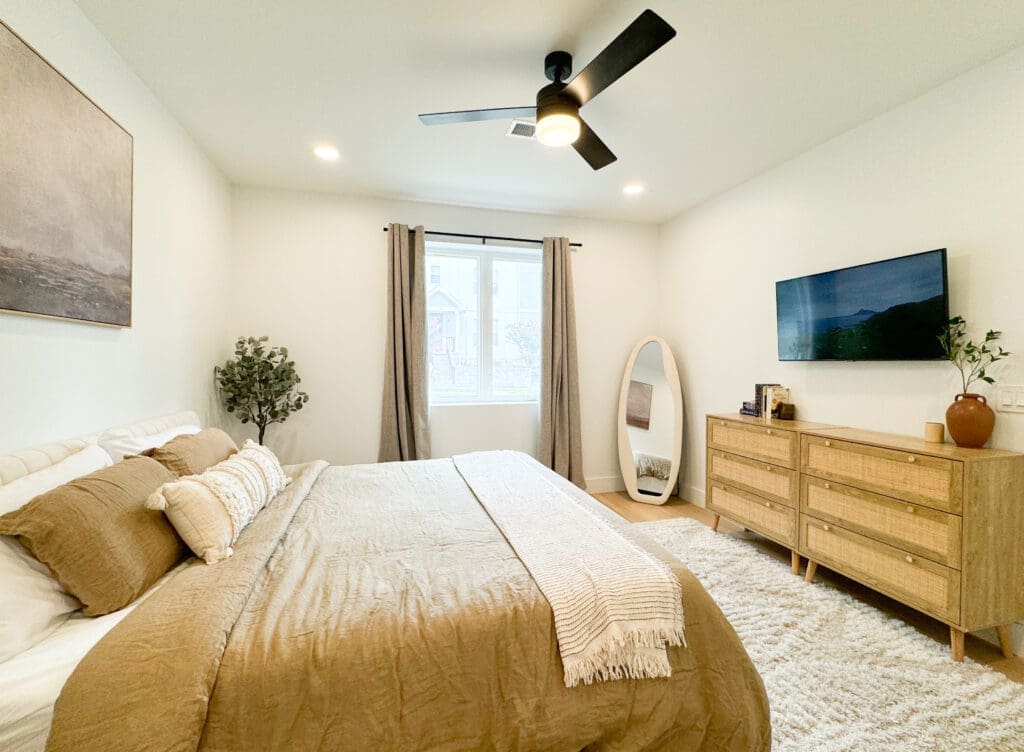 A bedroom with a bed, white walls, a ceiling fan, a dresser with a TV, a large mirror, and a window with curtains. The decor includes earthy tones and minimalistic furniture.