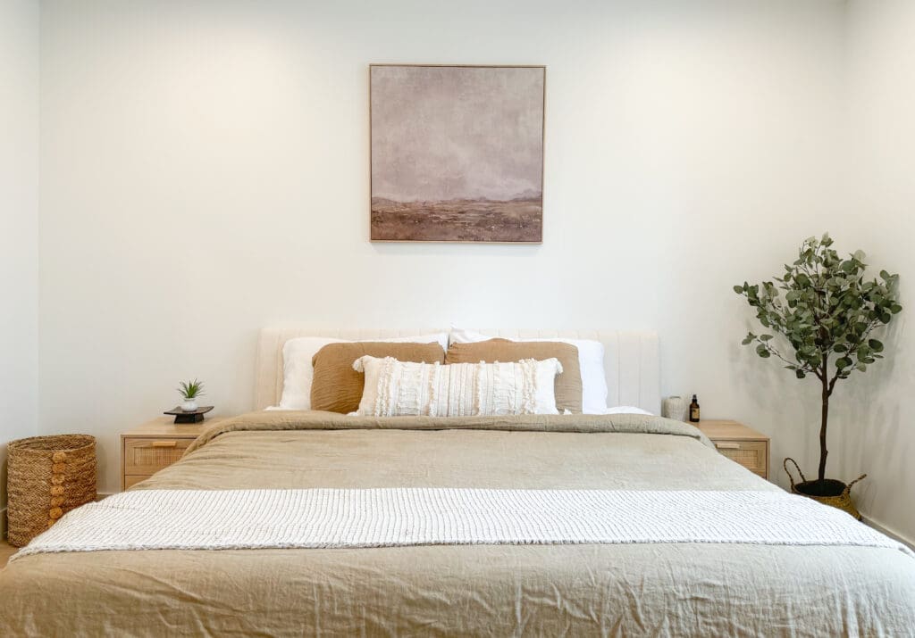 A neatly made bed with beige and white linens, large pillows, and a textured throw blanket. Flanked by nightstands with decor items, above the bed is a landscape painting. A plant stands to the side.