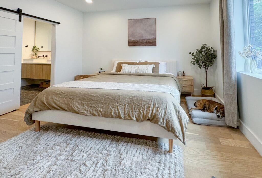 A modern bedroom with a neatly made bed, beige and white bedding, a large rug, plants, a canvas on the wall, and an open door leading to a bathroom. A dog is lying on a cushion near the window.