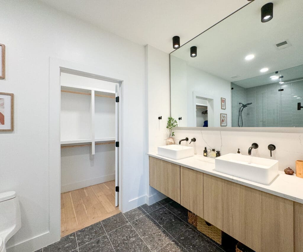 Modern bathroom with double sinks, large mirror, and light wood cabinets. View of adjacent walk-in closet with open shelving. Black fixtures, white walls, and a combination of tile and wood flooring.