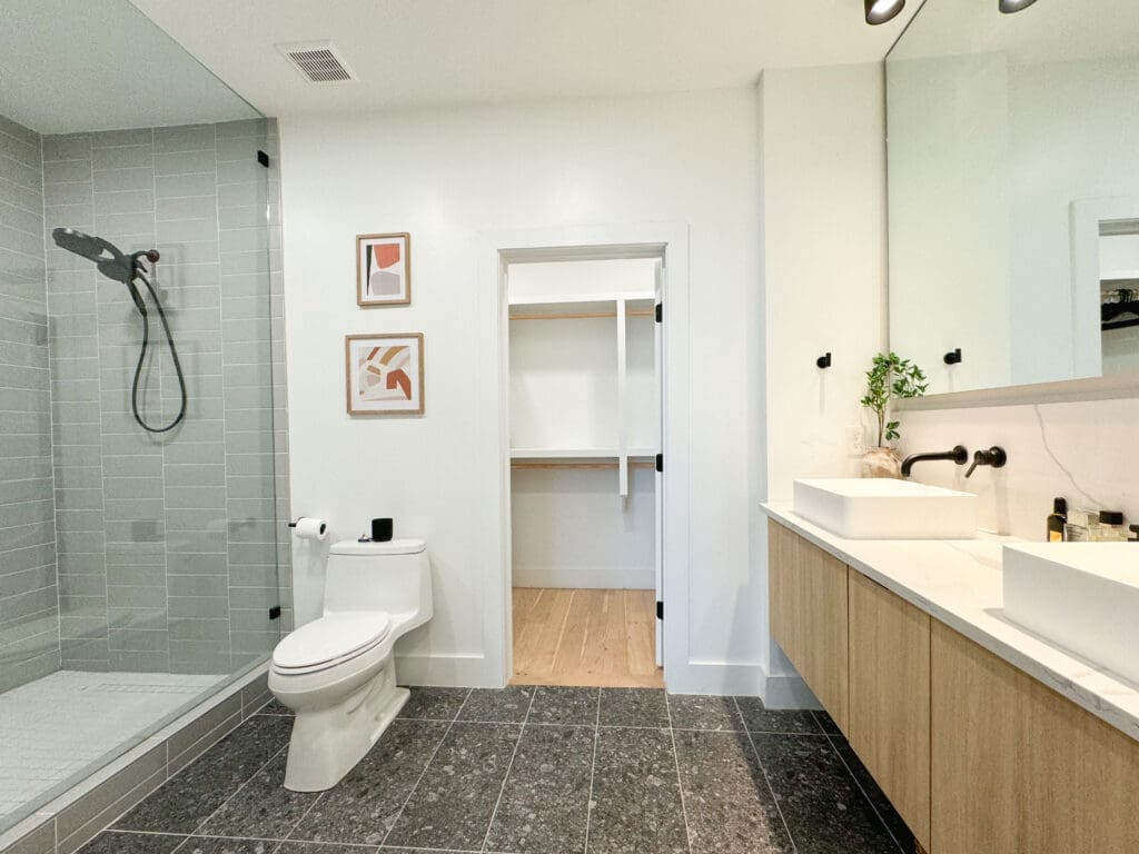 Modern bathroom featuring a walk-in shower with a glass door, a toilet, a double sink vanity with a large mirror, and artwork on the walls. A door leads to a walk-in closet.