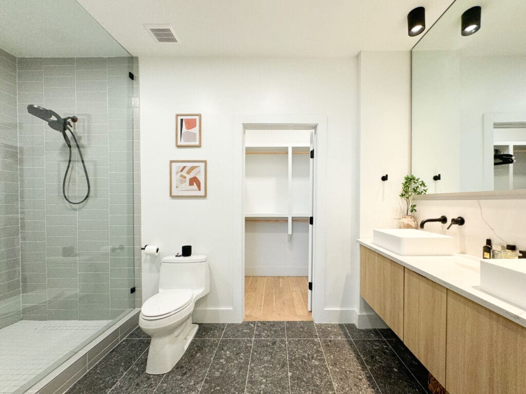 Modern bathroom with glass-enclosed shower, toilet, large mirror, double sink vanity, and walk-in closet. Walls are white with decorative art, and the flooring is dark tiled.