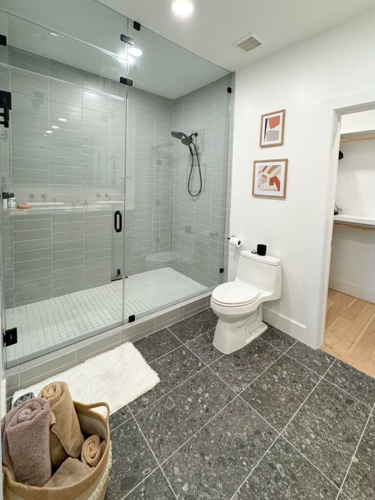 Modern bathroom with a glass-enclosed shower, gray tiled walls, and a white toilet. The floor has dark tiles. White walls feature two framed prints, and a door leads to another room. Towels in a basket.