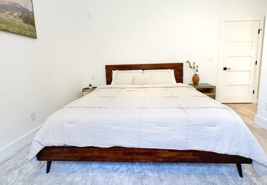 A minimalist bedroom features a king-sized bed with white linens, a wooden headboard, and side tables. A potted plant is on one table, and a closed white door is visible in the background.