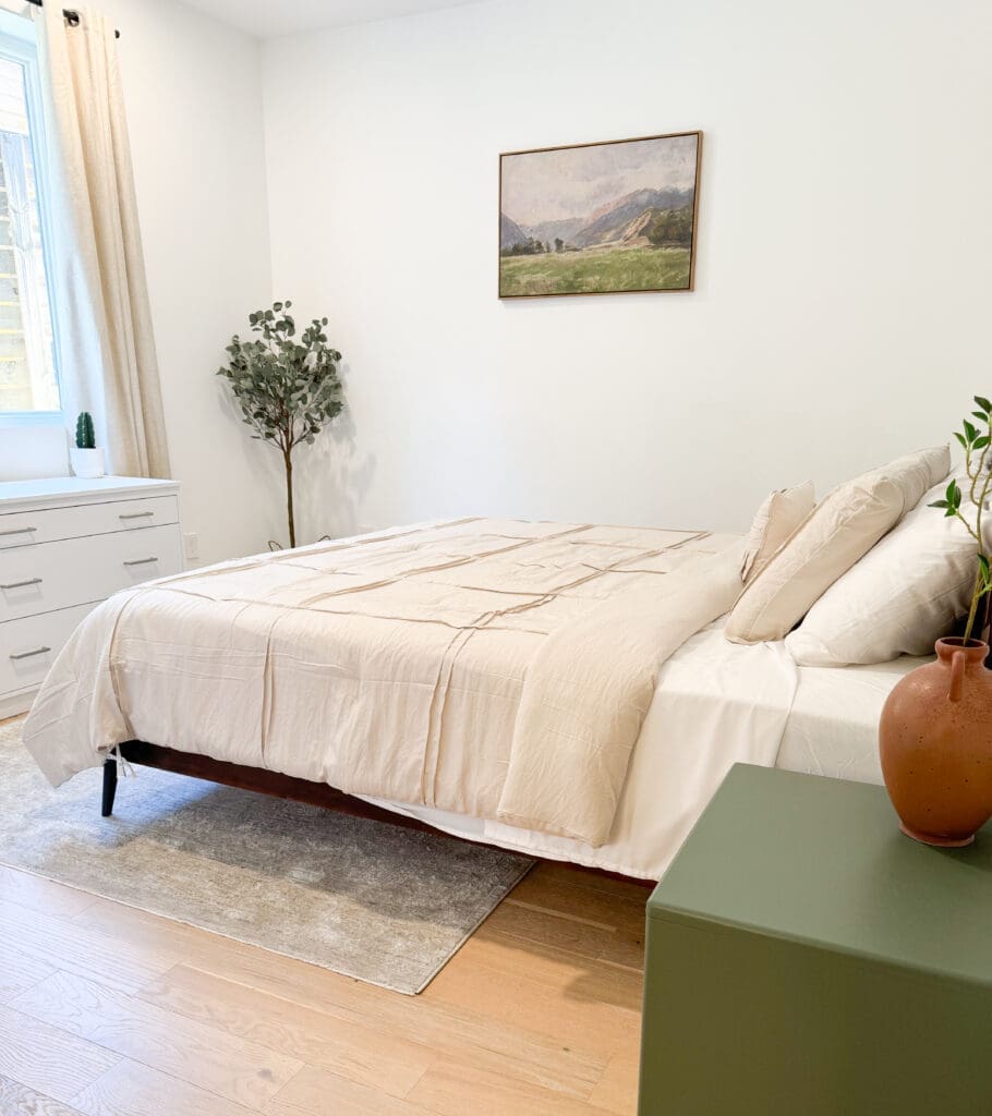 A neatly made bed with beige linens is centered in a minimalist bedroom. A dresser and potted plant are beside a window with light curtains. A landscape painting hangs on the white wall above the bed.