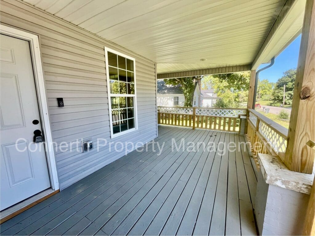 A covered wooden porch with gray flooring, beige siding, a white door, and a window. The porch has a wooden railing and overlooks a green yard with trees.