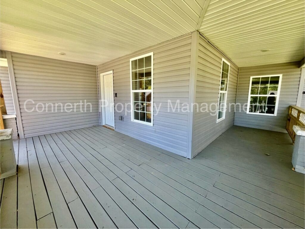 A spacious wooden deck with beige siding, two large windows, and a white door under a covered porch.