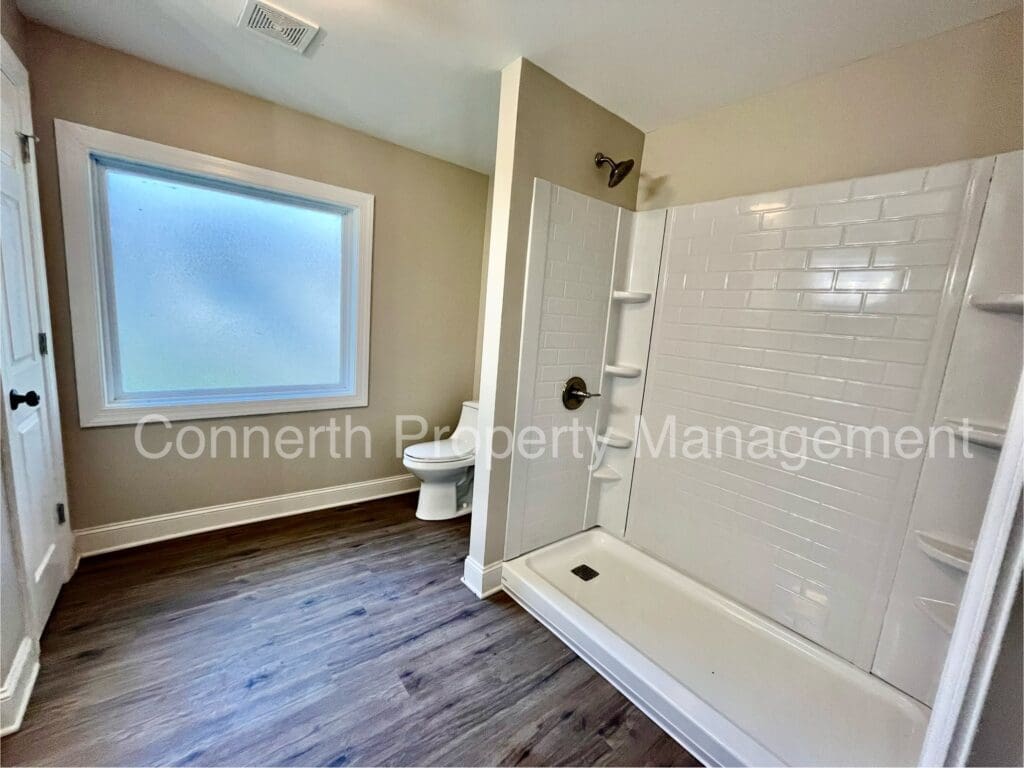 Bathroom with a large walk-in shower, a toilet, and a frosted window. Wood-style flooring and light-colored walls.