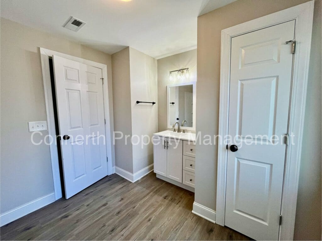 A bathroom with light wood flooring, white vanity with a mirror and sink, and two white doors. One door is open, revealing a closet. The walls are painted a light beige color.