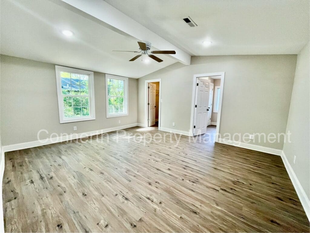 Spacious empty room with wood flooring, beige walls, and a ceiling fan. Two large windows let in natural light. Doorway leads to another area. Text overlay reads: 