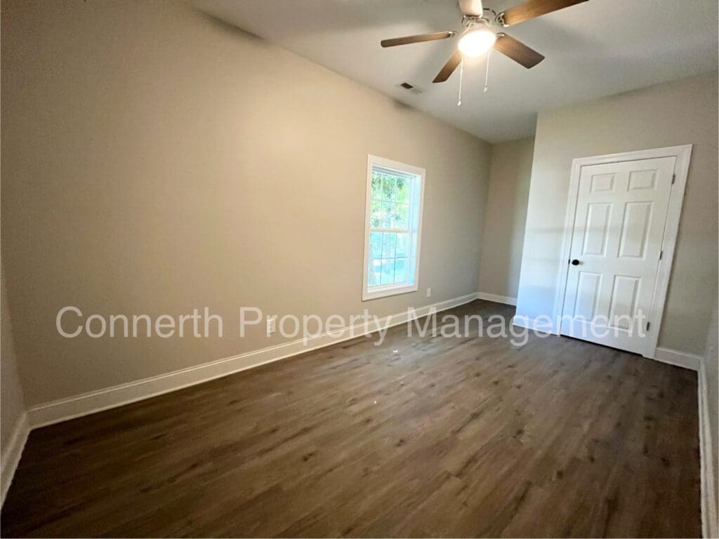 Empty room with wood flooring, beige walls, a window, a ceiling fan, and a closed white door.