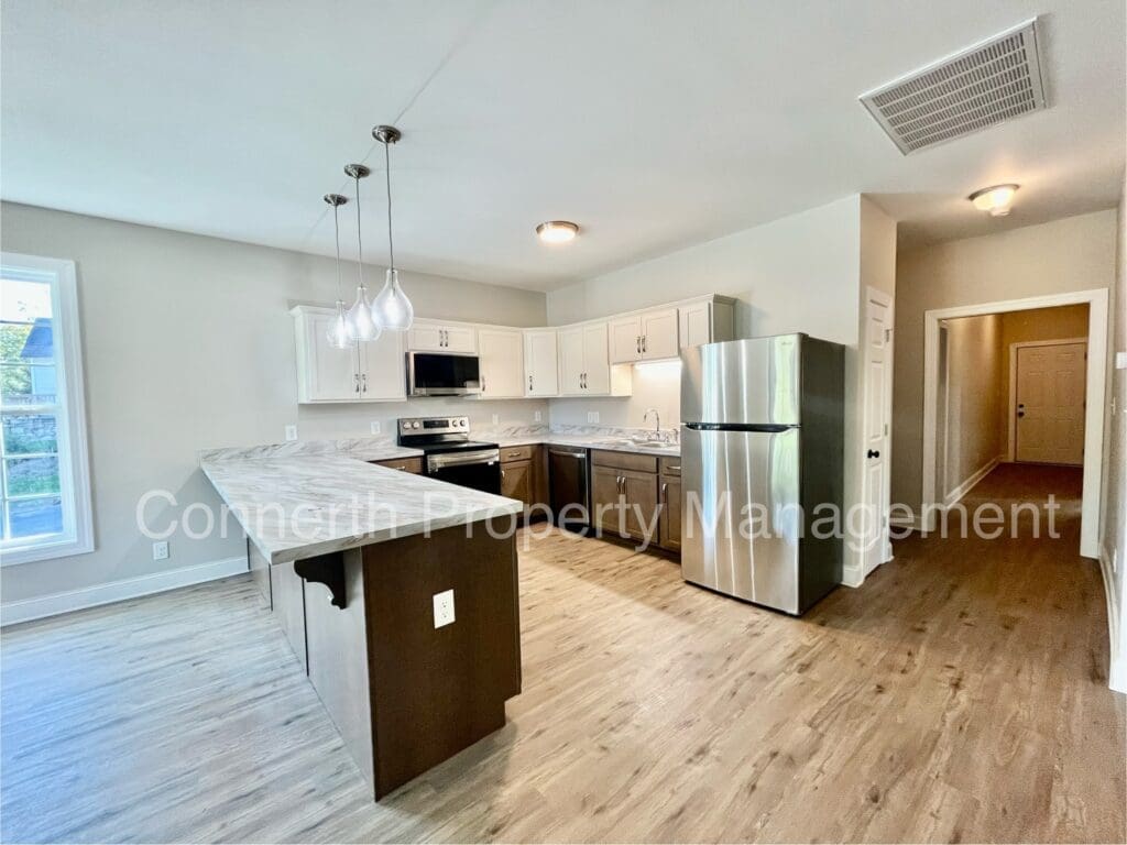 Modern kitchen with stainless steel appliances, white cabinets, and a large island. Wood flooring and pendant lights add to the neutral decor.